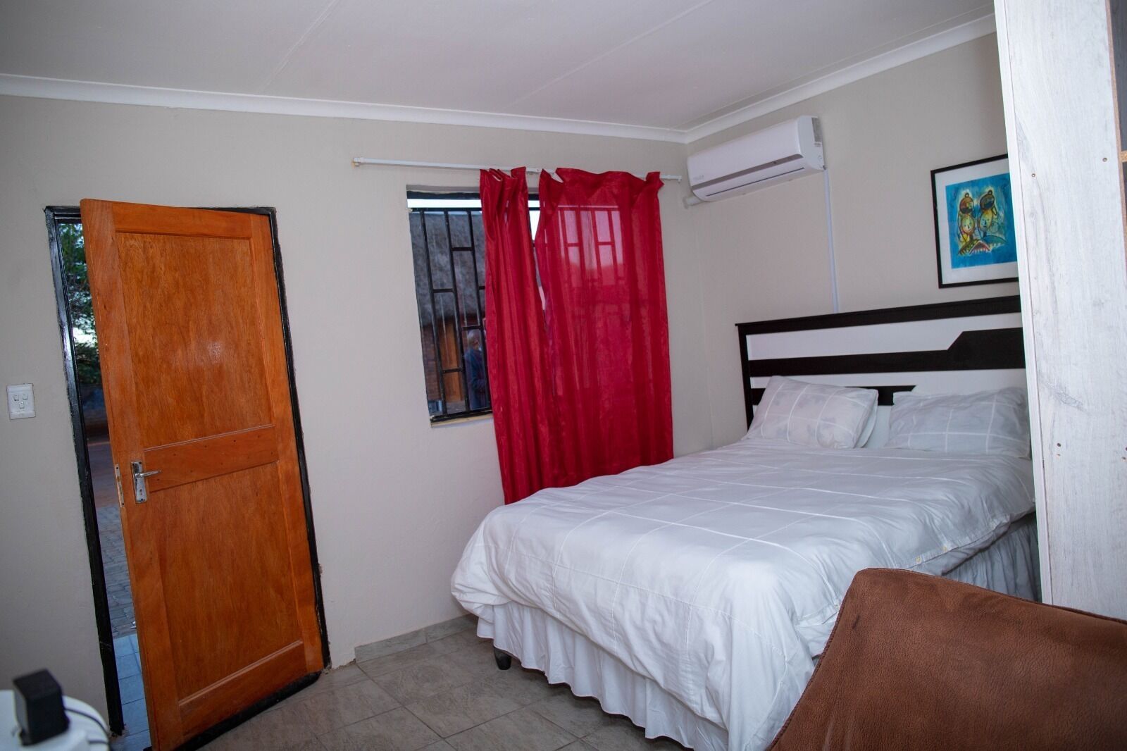 A bedroom with a bed, a window with red curtains, a door, and an air conditioning unit.