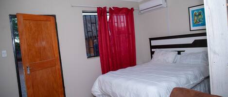 A bedroom with a bed, a window with red curtains, a door, and an air conditioning unit.