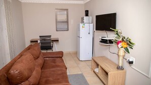 Una sala de estar con un sofá marrón, un refrigerador blanco, un televisor de pantalla plana, una mesita de madera y un jarrón con flores.