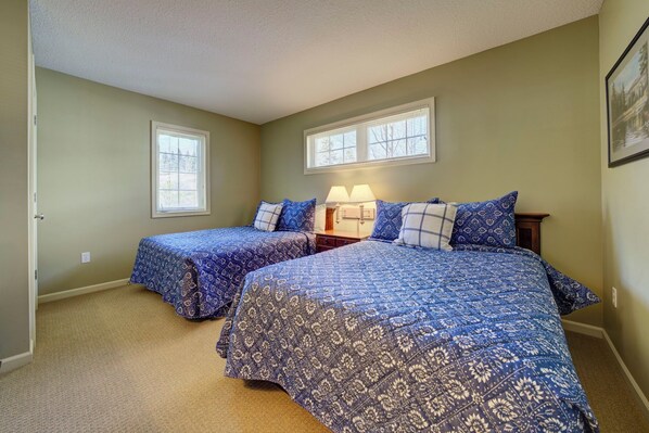 A bedroom with two beds, a nightstand, a lamp, and two windows.