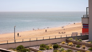 Une vue sur la plage avec une rambarde, des personnes se promenant sur la plage et un bâtiment dont une partie est rouge et l’autre bleue.