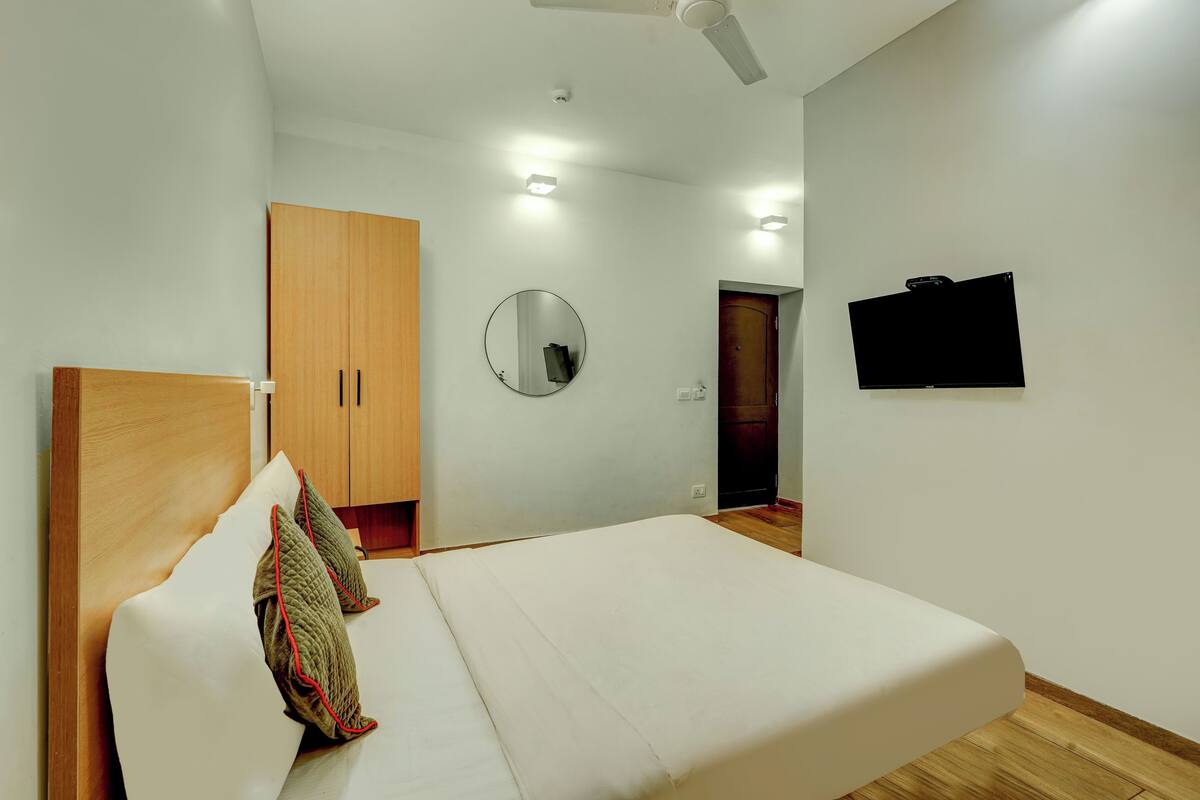 A bedroom with a bed, wooden wardrobe, a round mirror, a television, and a ceiling fan.