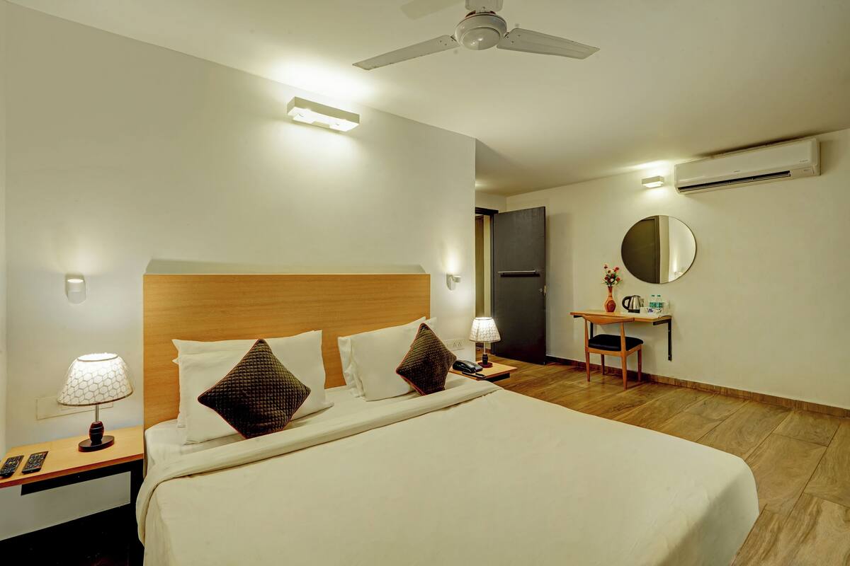 A hotel room with a large bed, bedside lamps, a desk with a chair, a mirror, and an air conditioning unit.