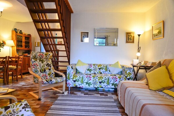 A cozy living room with a wooden staircase, a floral sofa, a patterned armchair, and a dining area with wooden furniture.