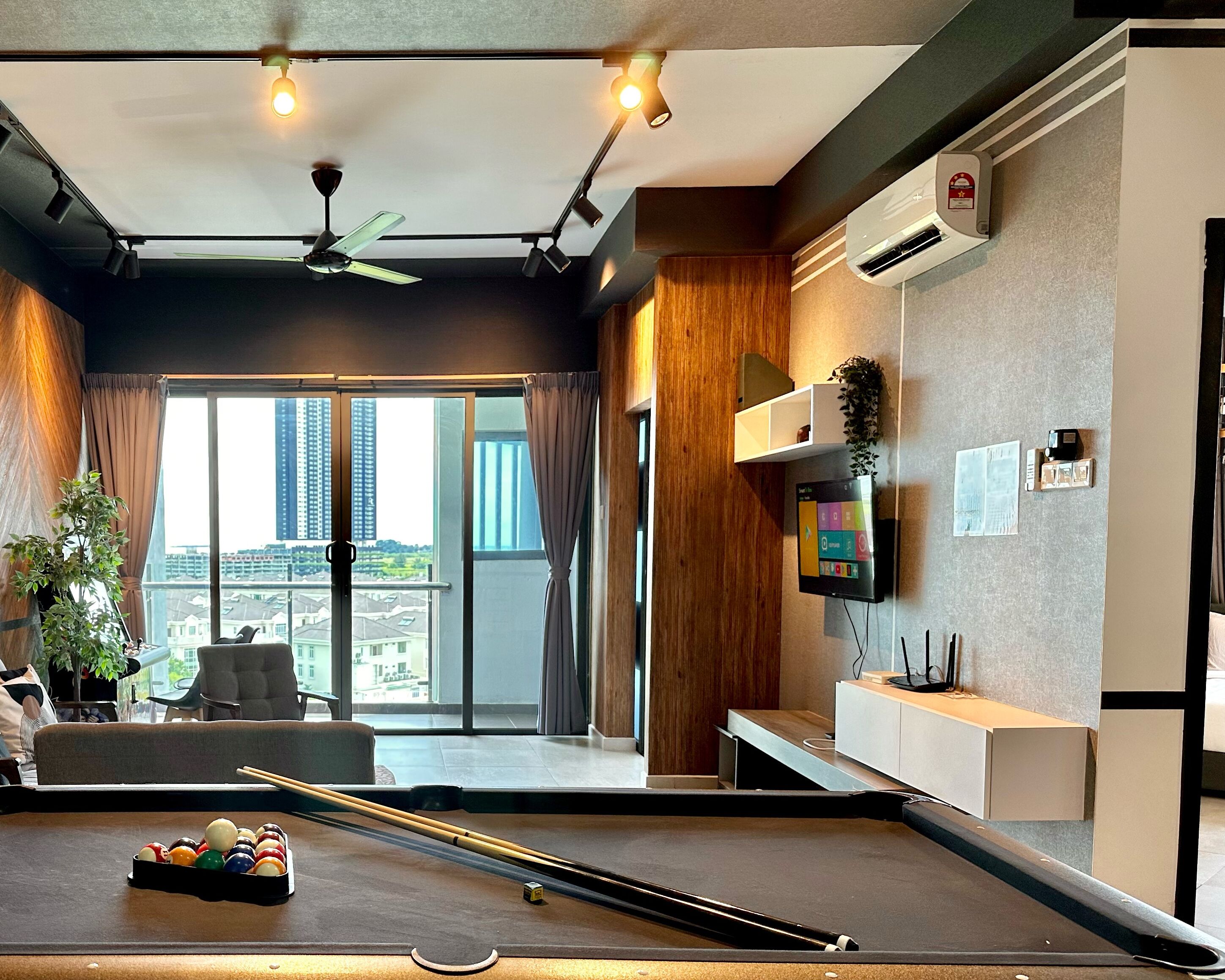 Una habitación de hotel moderna con mesa de billar, ventilador de techo, un ventanal con cortinas y un televisor montado.