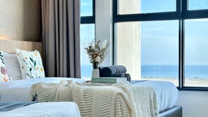 A bedroom with a large window, a bed with white bedding, a decorative plant, and a book titled "NORDIC INTERIORS HOMES." - Atlantis Residence 2 (Malacca City)