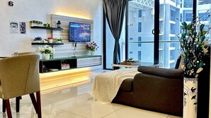 A modern living room with a sofa, a TV, a ceiling fan, and a large window with a view of buildings. - Atlantis Residence 2 (Malacca City)