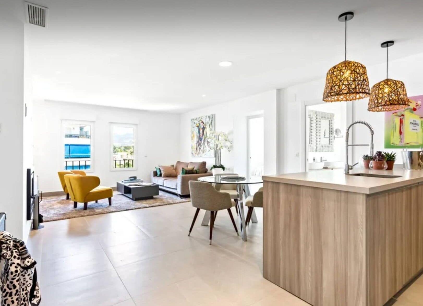 Uma sala de estar moderna com sofá, mesa de centro e cadeiras amarelas. Há uma área de jantar com mesa e cadeiras, e uma cozinha com bancada e luminárias pendentes.
