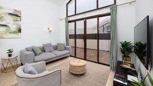 A modern living room with a grey sofa, a round ottoman, a flat-screen TV, and a large window with curtains. - Villa Cindy - Three Bedroom Villa, Sleeps 6 (Playa Blanca)