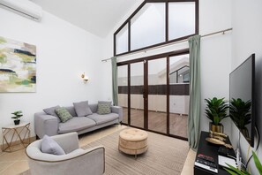 Un salon moderne avec un canapé gris, un pouf rond, un téléviseur à écran plat et une grande fenêtre avec des rideaux.