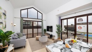 A modern living room with a sofa, a dining table set for two, and a large sliding glass door leading to an outdoor area with a pool. - Villa Cindy - Three Bedroom Villa, Sleeps 6 (Playa Blanca)