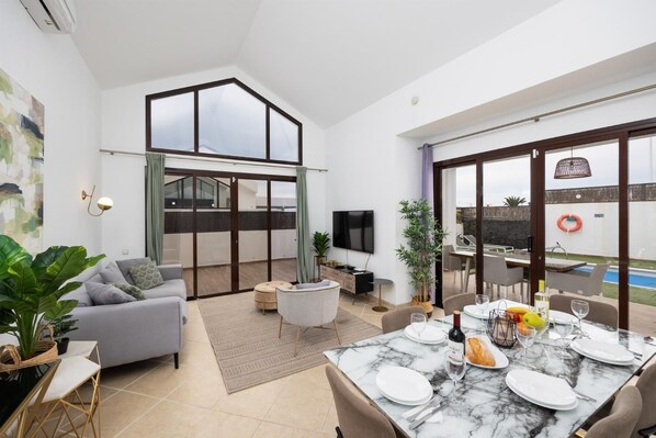 Un salon moderne avec un canapé, une table à manger pour deux personnes et une grande porte-fenêtre coulissante donnant sur un espace extérieur avec une piscine.