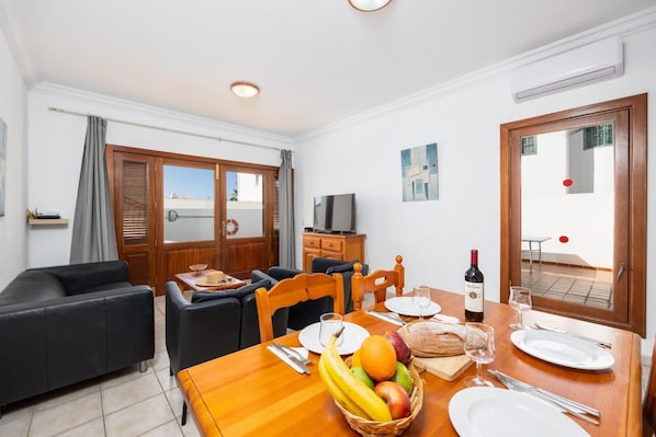Una sala moderna con un sofá, una mesa de comedor dispuesta con fruta, vino y platos, y un televisor sobre un mueble de madera.