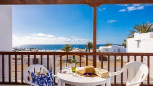 Un balcon avec vue sur la mer, des chaises blanches, une table avec une bouteille, des verres et un chapeau.