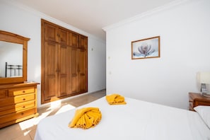 A bedroom with a large wooden wardrobe, a bed with white bedding, a bedside table with a lamp, and a framed picture on the wall.
