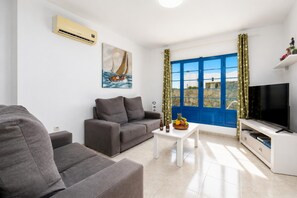 A modern living room with a grey sofa, a white coffee table with fruit, a flat-screen TV, and a window with blue shutters.