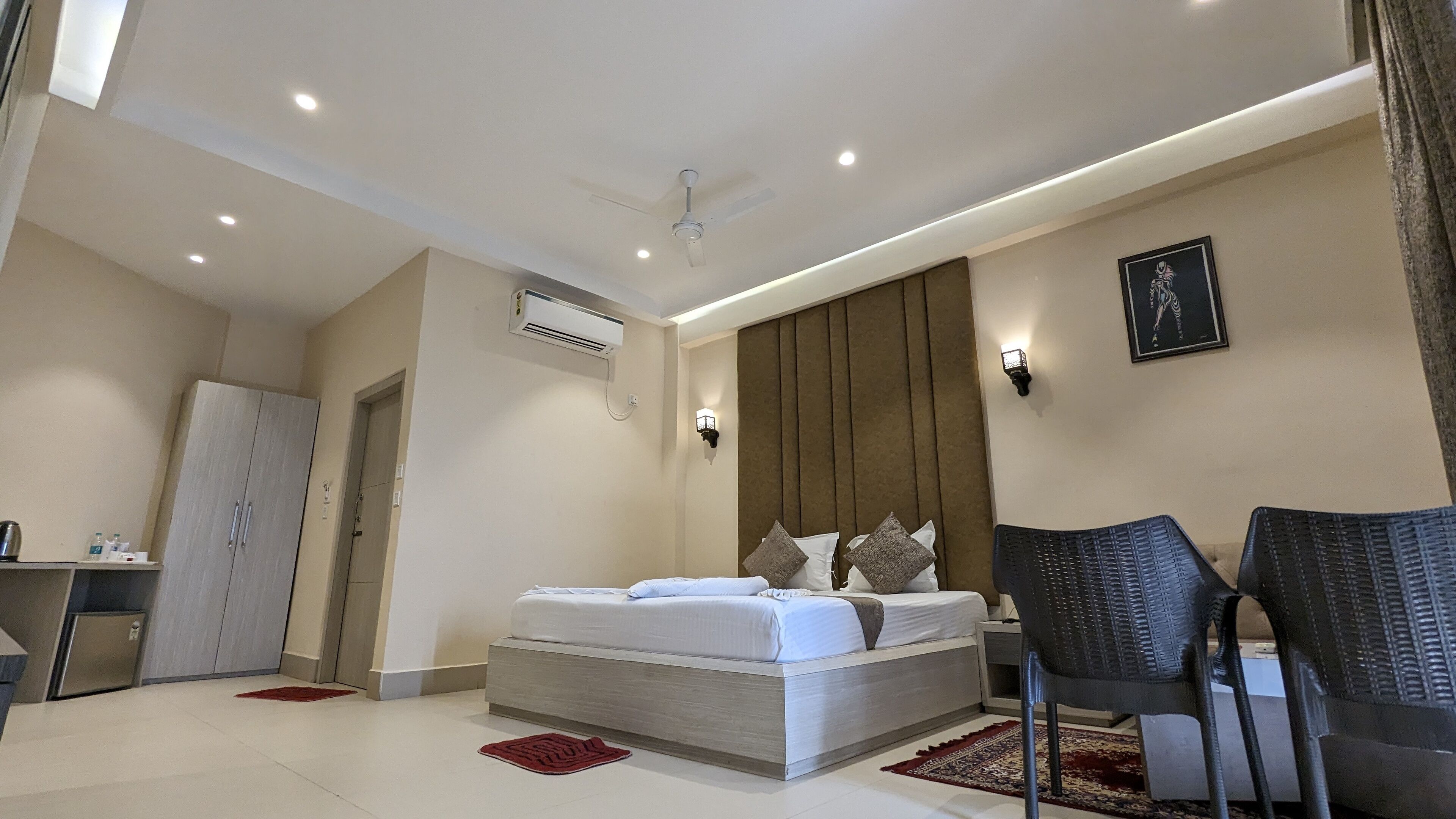A modern bedroom with a large bed, a desk, and a chair. There is a wardrobe, a ceiling fan, and a wall-mounted air conditioner.