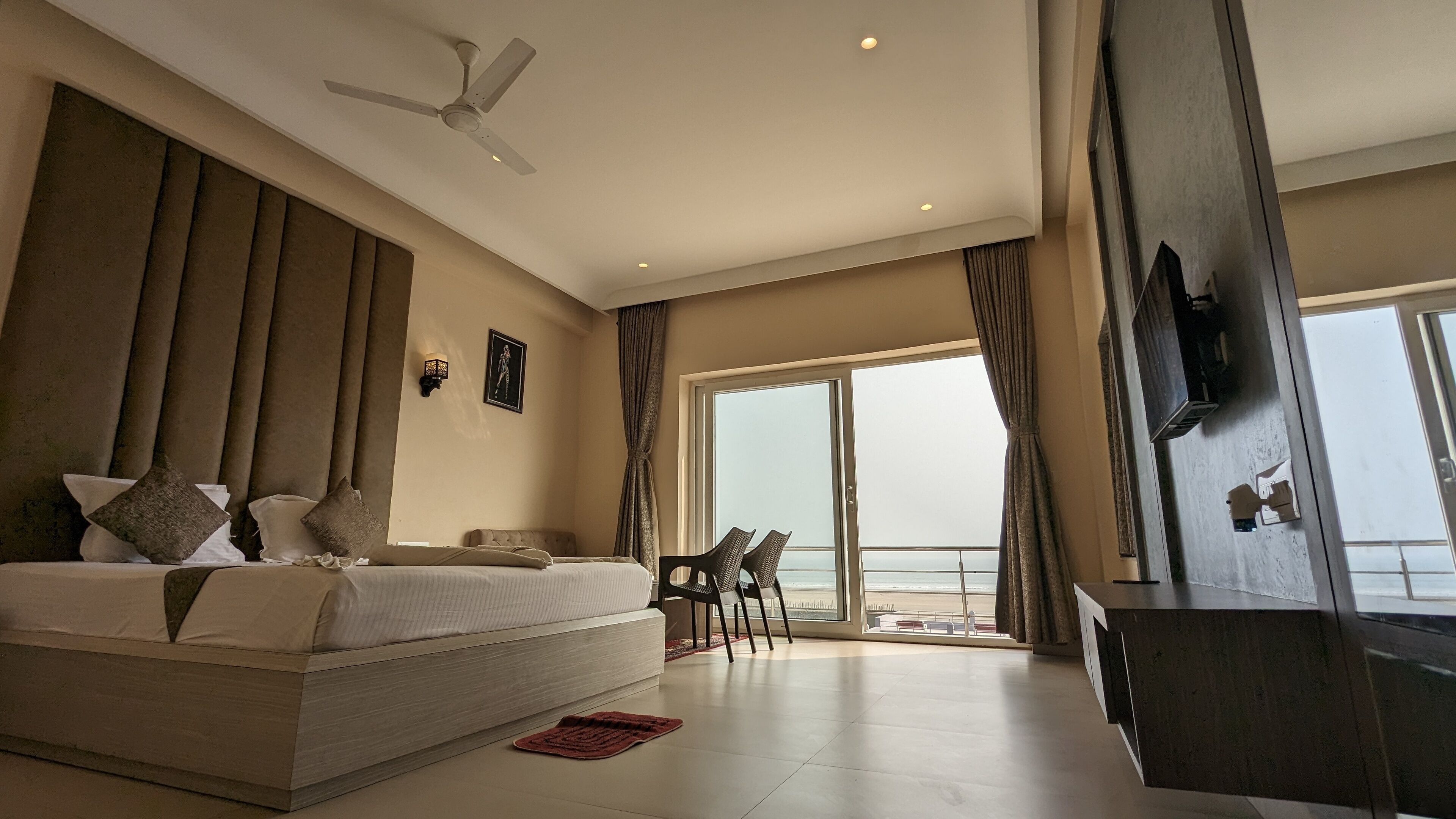 A modern hotel room with a large bed, a ceiling fan, a TV mounted on the wall, and a balcony with a view.