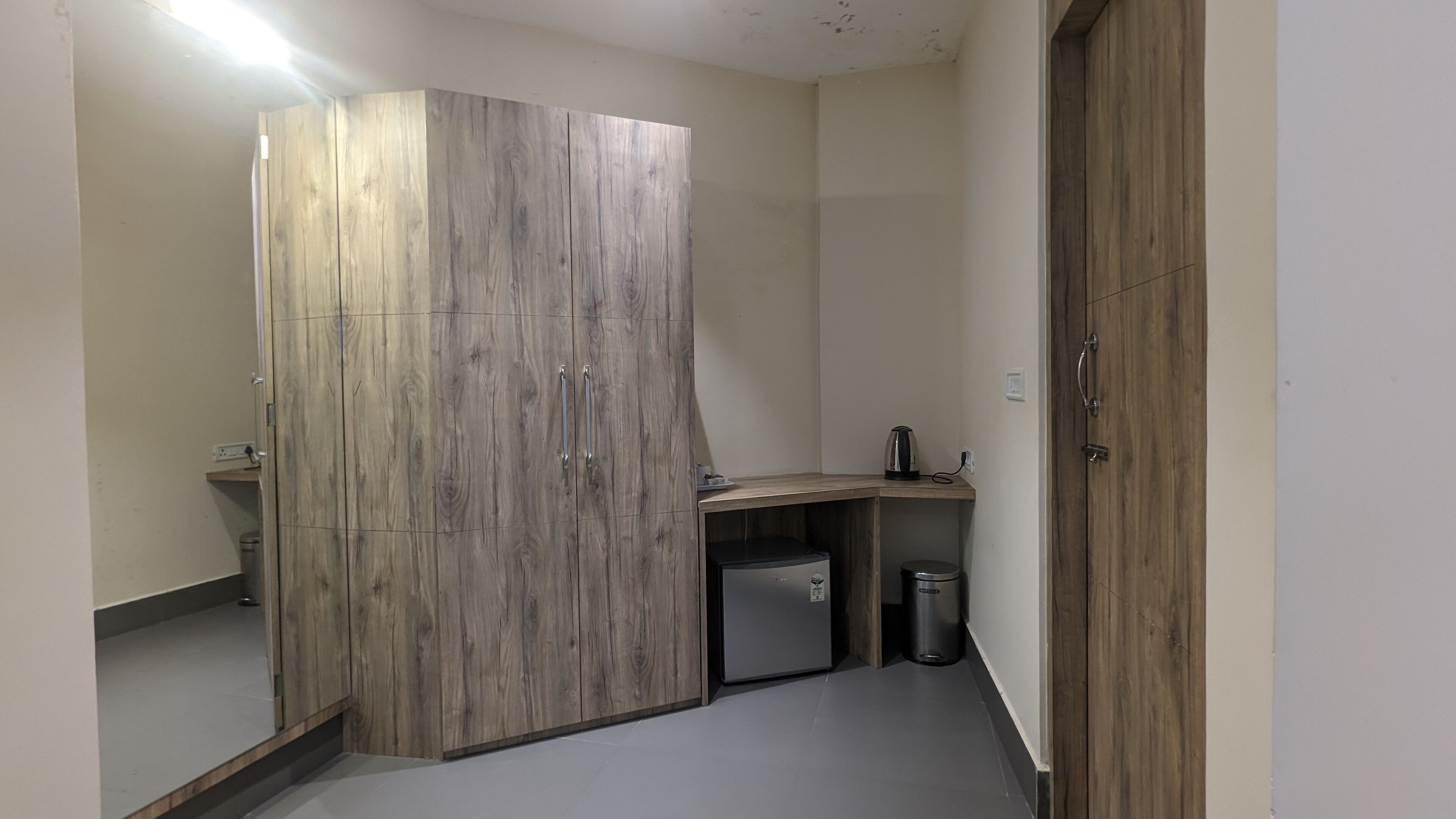 A compact bathroom with a wooden vanity, a small sink, a kettle, and a trash bin.