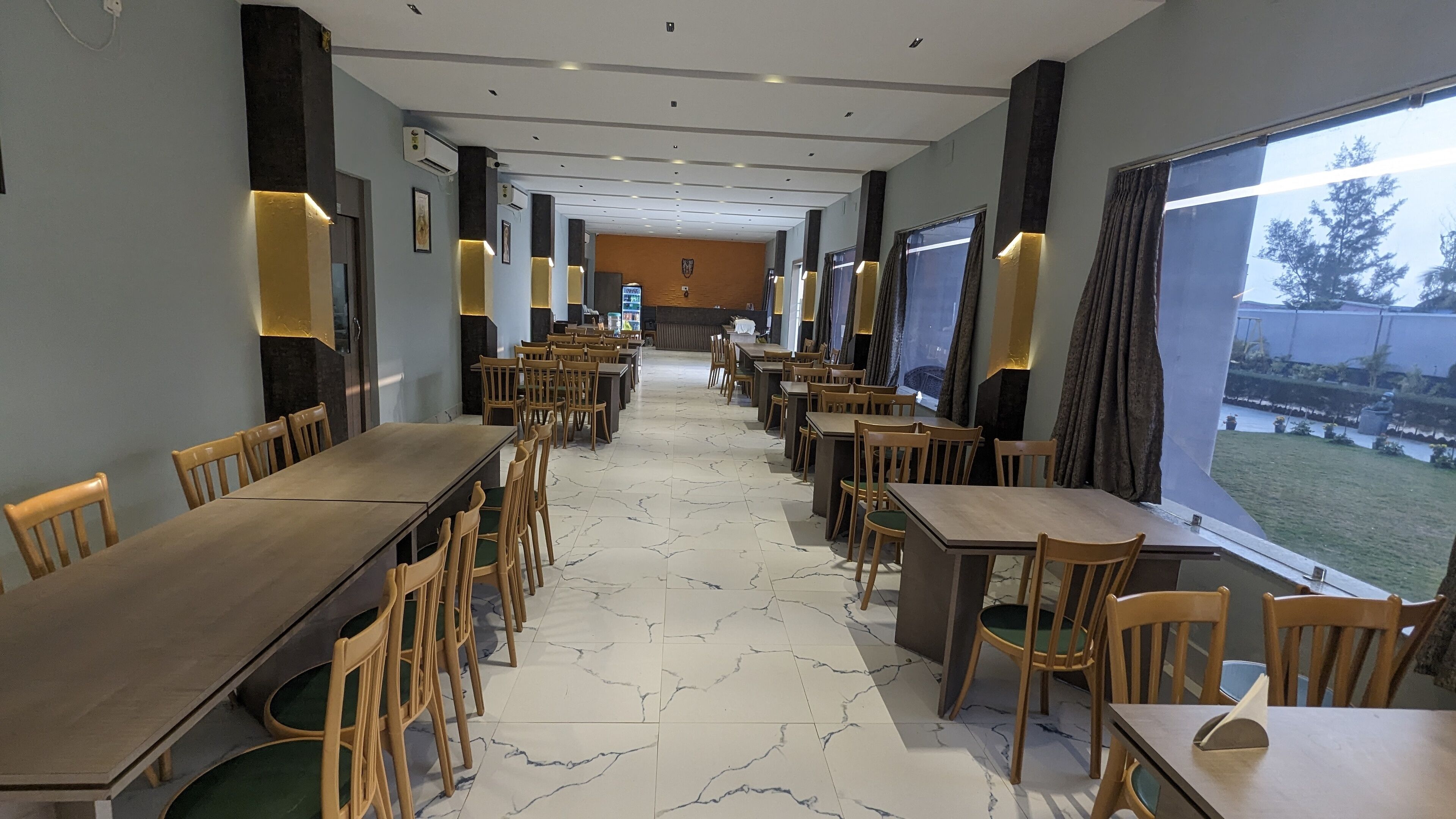 A spacious dining area with long tables and chairs, a marble floor, and large windows offering a view of the outdoors.