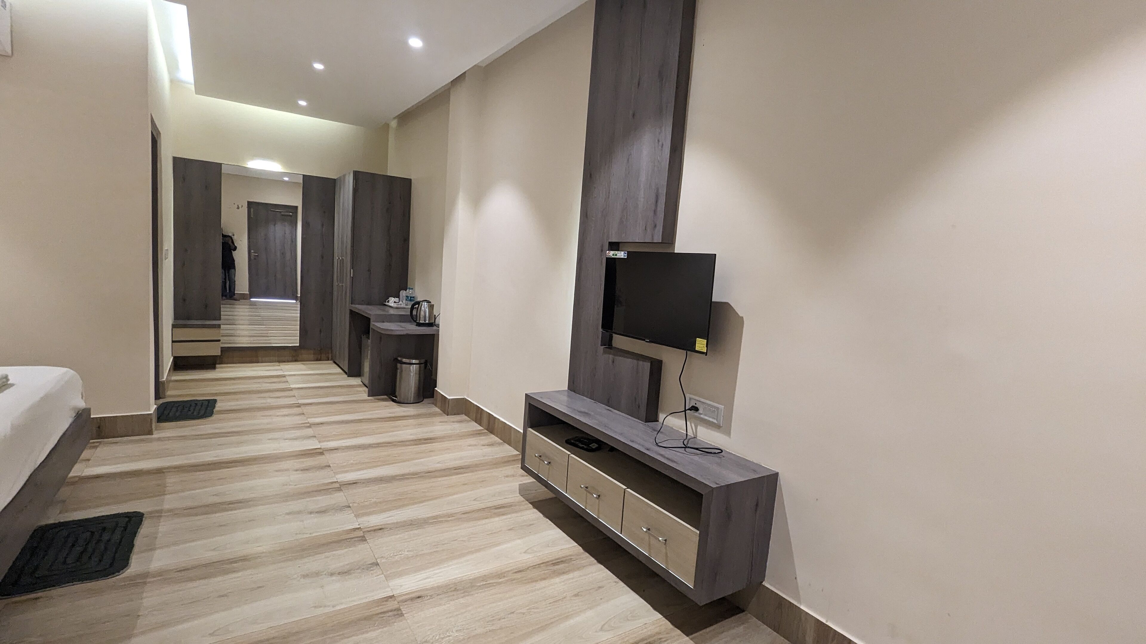Une chambre d’hôtel moderne avec un plancher en bois, un téléviseur à écran plat fixé sur une console en bois, une petite table avec une bouilloire et un miroir qui reflète la pièce.