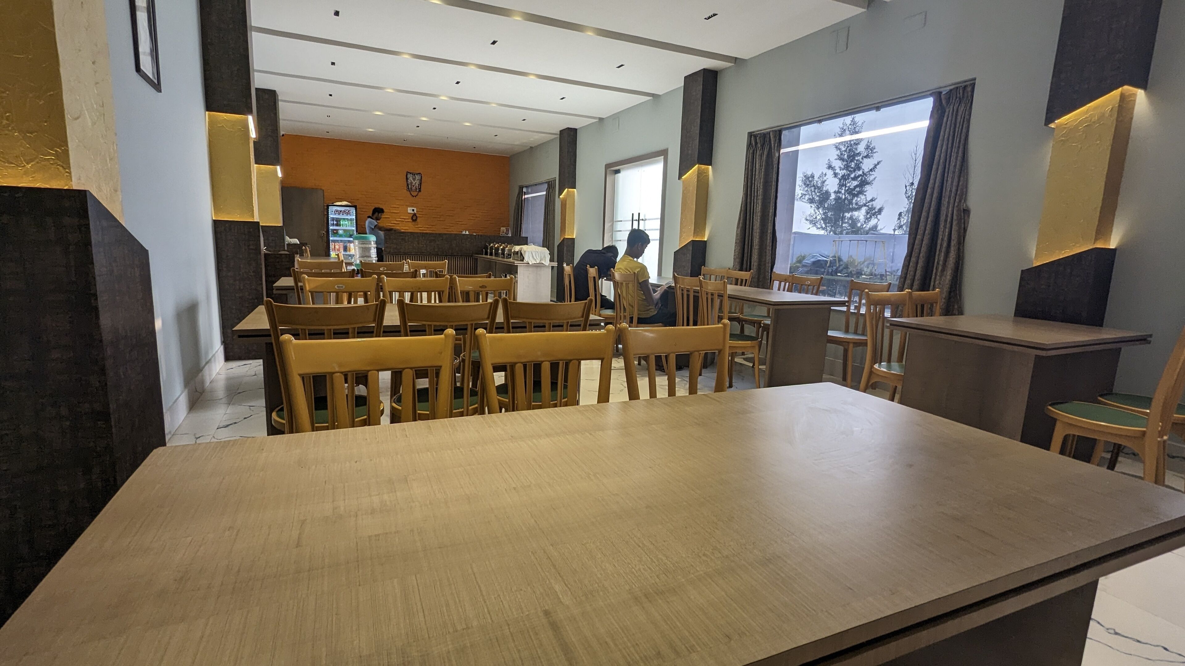 A dining area with wooden tables and chairs, a Coca-Cola refrigerator, and a view of the outdoors through large windows.