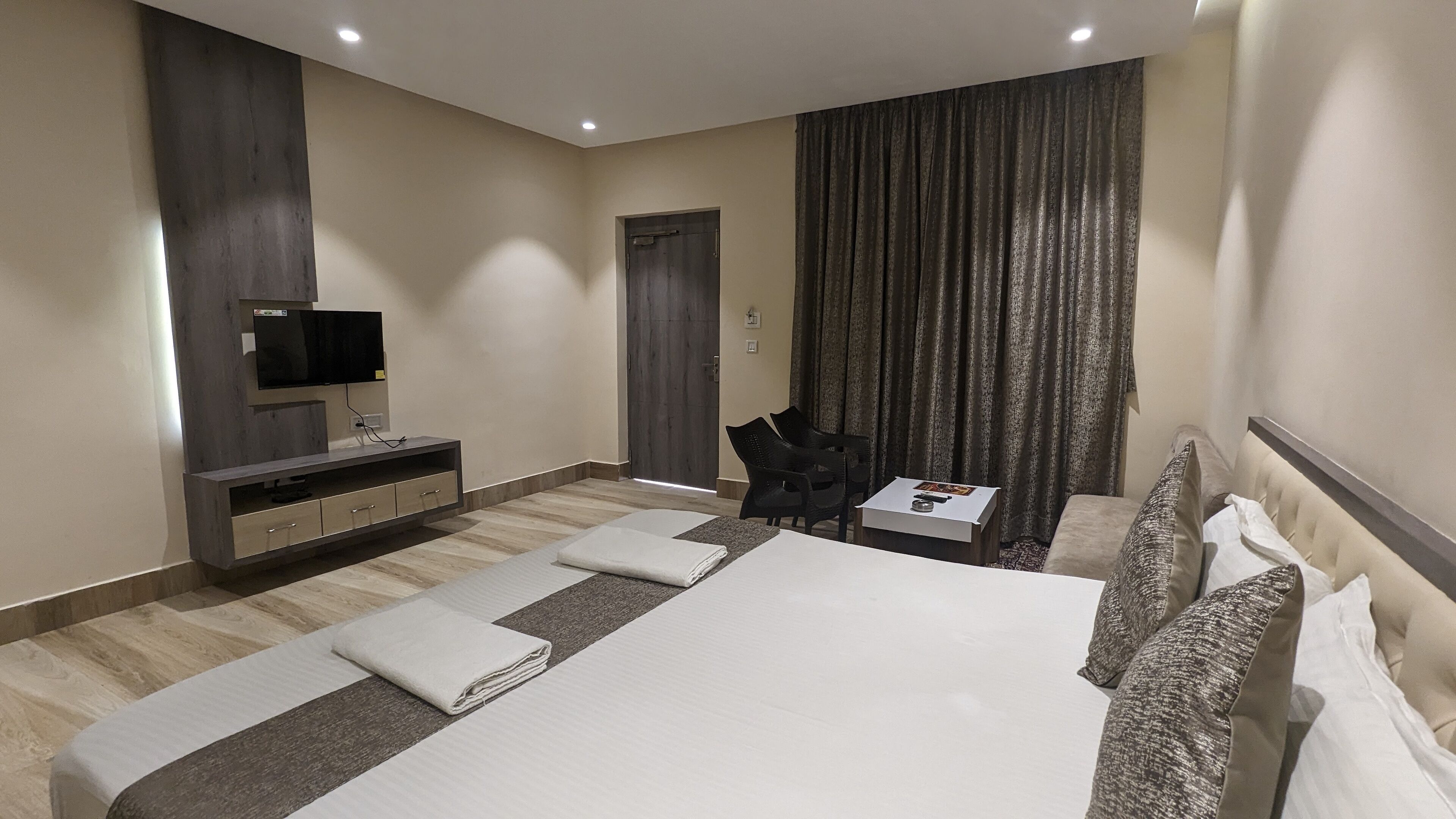 A modern hotel room with a large bed, a flat-screen TV, a desk with a chair, and a dark curtain.