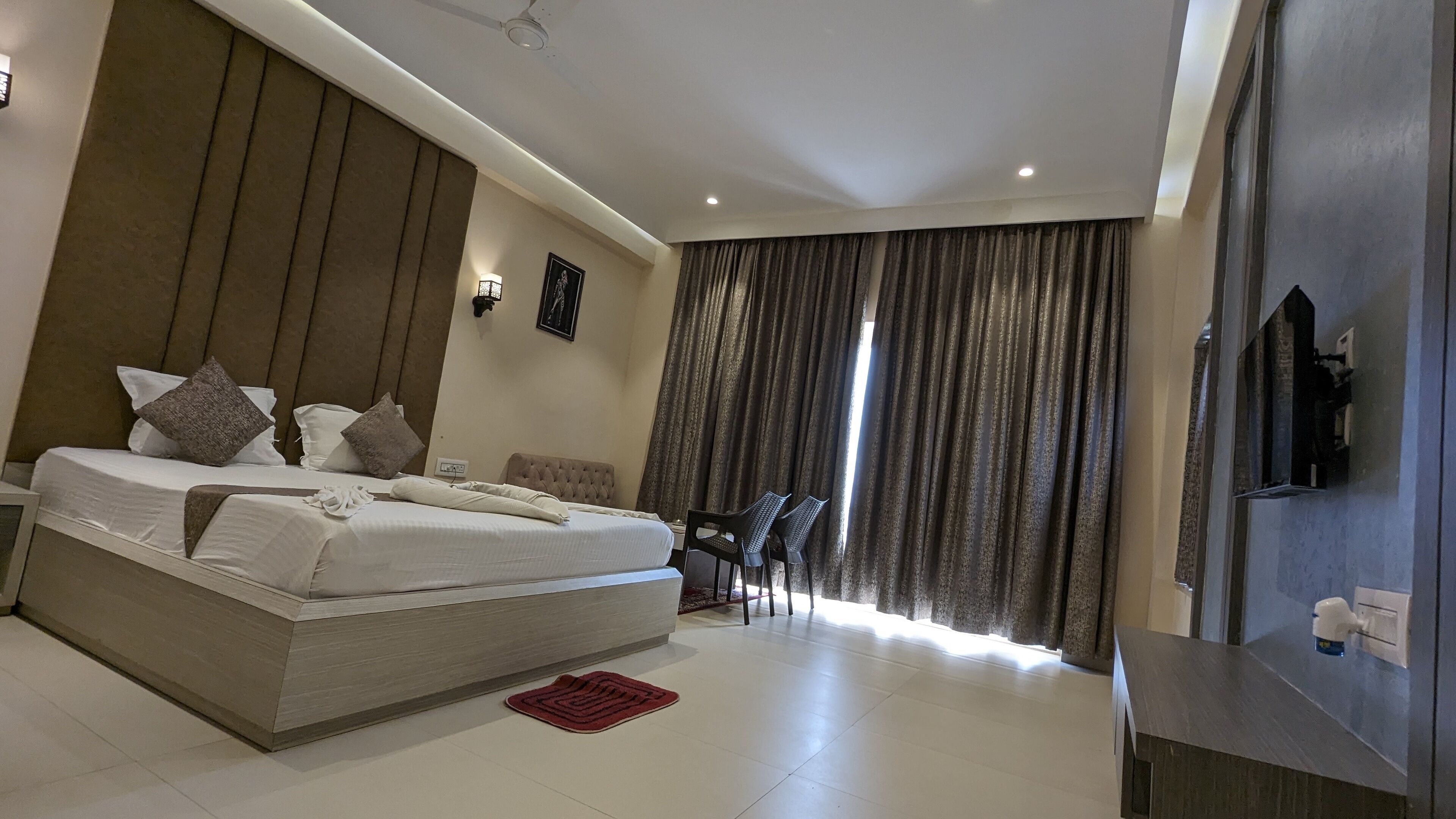 A modern hotel room with a large bed, a desk with chairs, a TV, and a view of the outdoors through curtains.