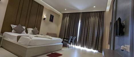 A modern hotel room with a large bed, a desk with chairs, a TV, and a view of the outdoors through curtains.