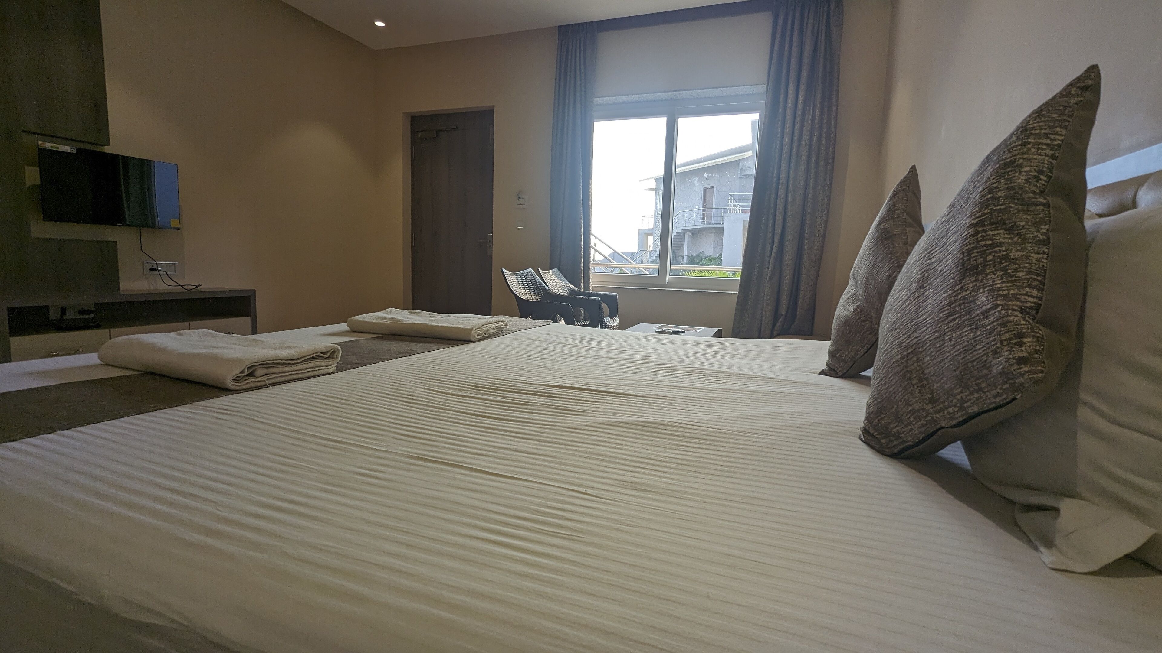 A neatly made bed with pillows and a folded towel, a chair, a television mounted on the wall, and a window with curtains.