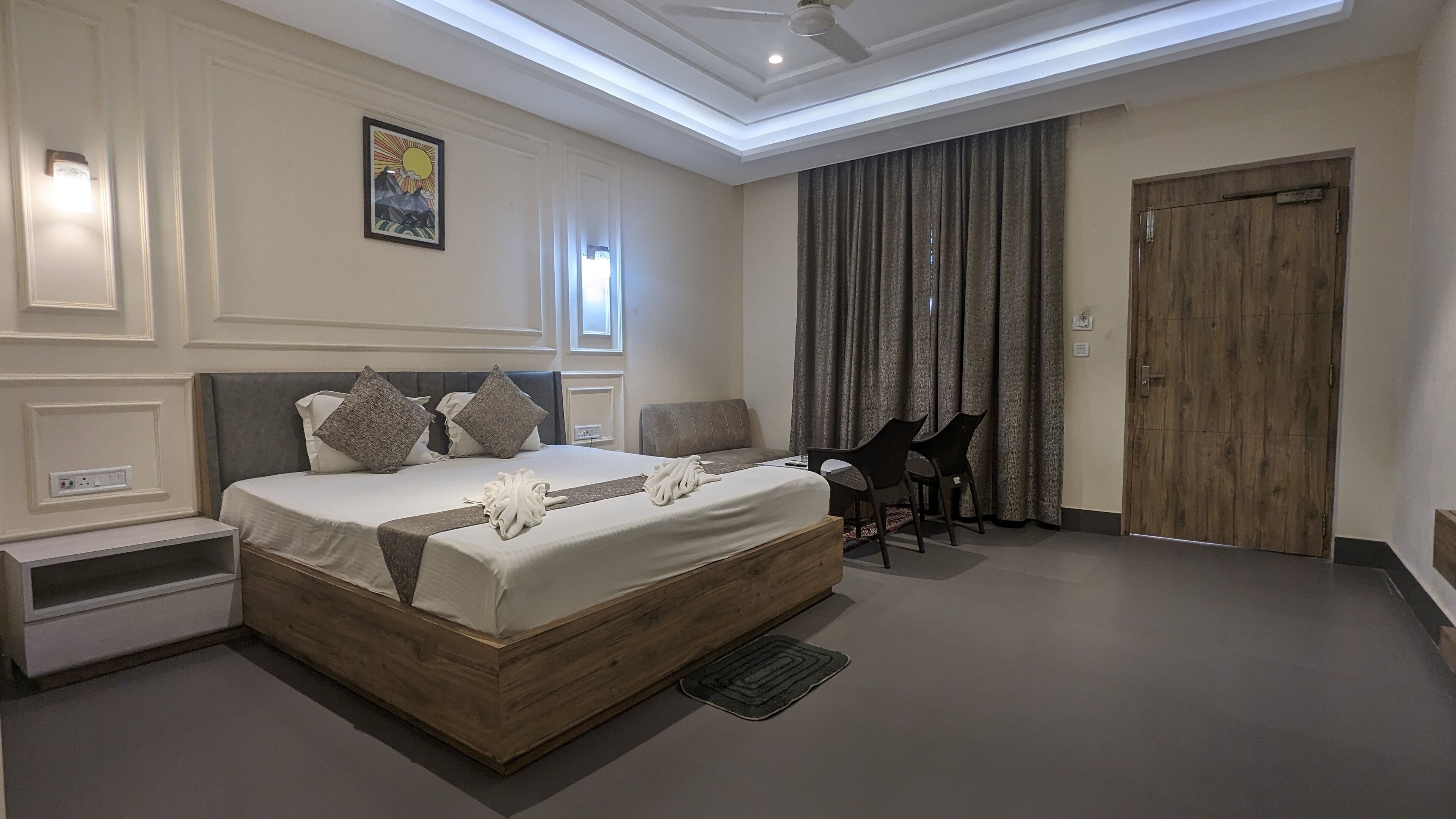 Un dormitorio moderno con una cama grande, mesitas de noche, un escritorio con sillas, un ventilador de techo y una puerta de madera.