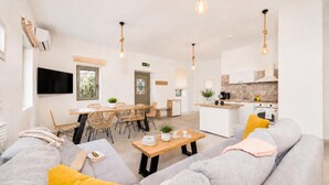 A modern living room with a sofa, a dining area with chairs, a kitchen with appliances, and pendant lights.