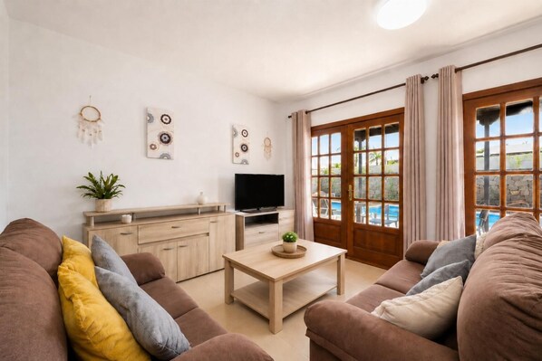 A living room with a TV, wooden furniture, a plant, and a view of a swimming pool.