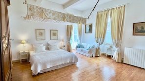 Een slaapkamer met een groot bed, een nachtkastje met een lamp, een stoel, een klein tafeltje en een raam met gordijnen.