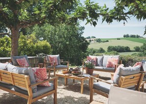 An outdoor seating area with wooden furniture and cushions, surrounded by greenery and a view of the countryside.