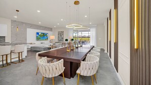 A spacious kitchen with a marble backsplash, a dining area with a long wooden table and white upholstered chairs, and a large window offering a view of the outdoors. - Fabulous Fun 8 Bed Pool Home with Game Room-4297LA (Davenport)