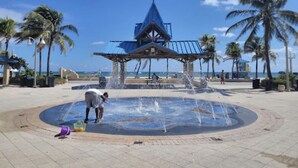 Une fontaine publique avec des jets d’eau, entourée de palmiers et d’un pavillon au toit bleu.