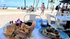 Une table à manger dressée avec des plats de fruits de mer, donnant sur la plage, avec des palmiers et des personnes en arrière-plan.