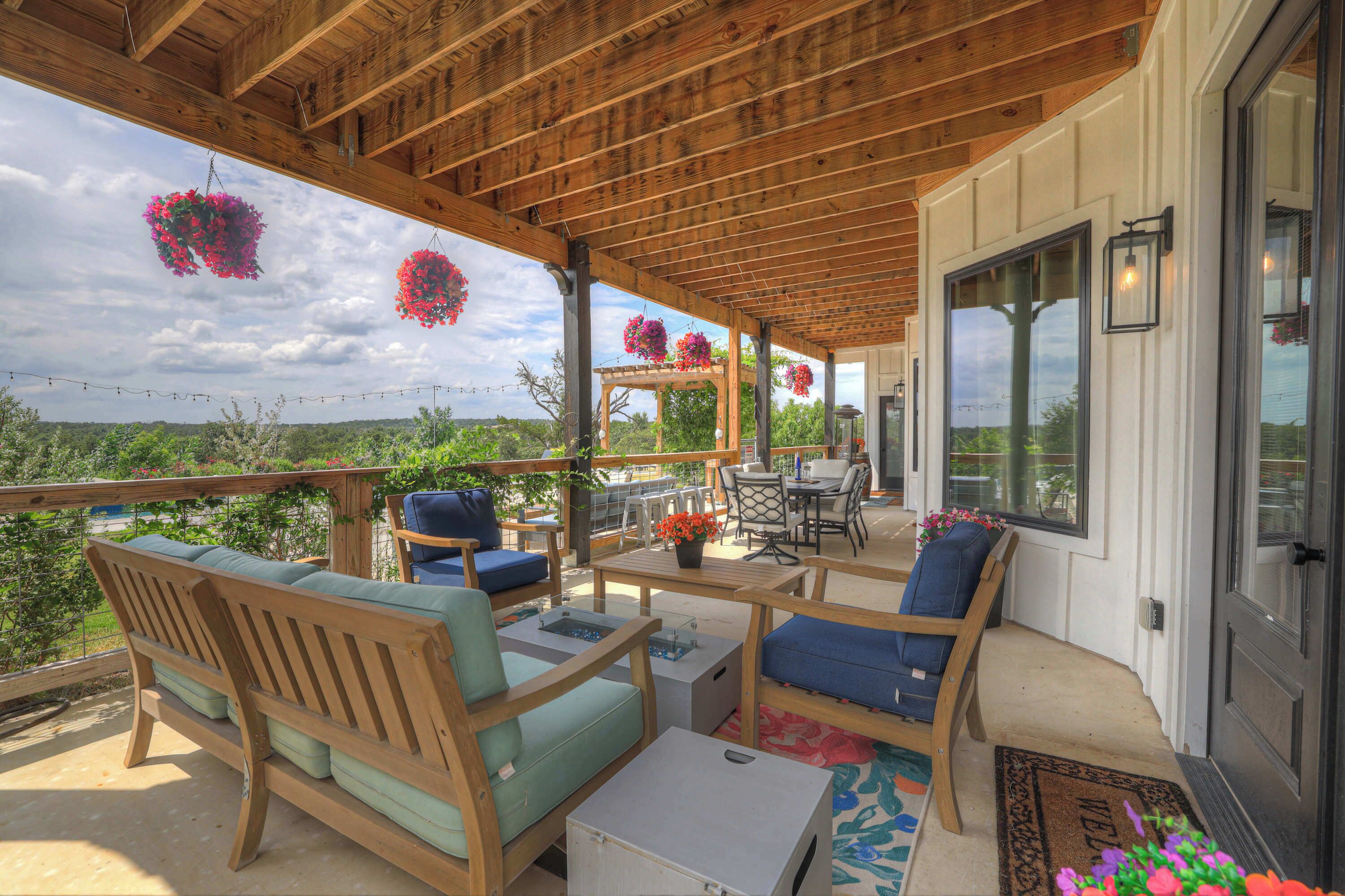 A covered patio with wooden furniture, a pergola with hanging flower baskets, and a view of the landscape.