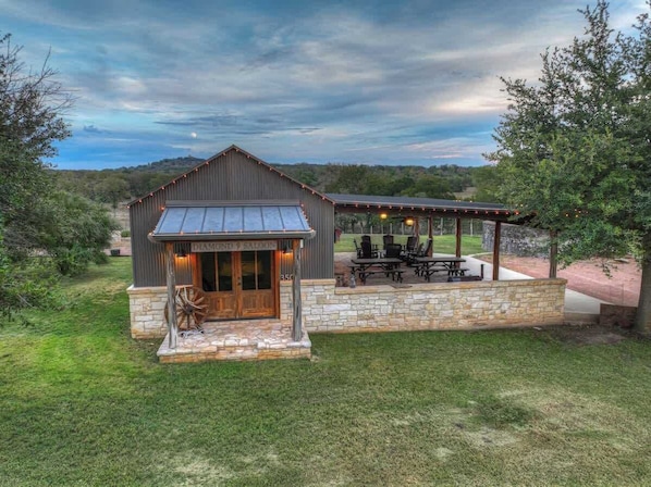 A rustic building with a covered outdoor seating area and a stone foundation. - Coco at Sunset Valley Ranch (Fredericksburg)