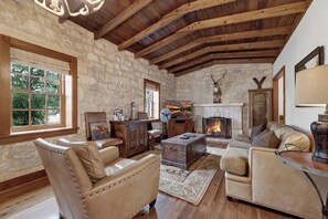 A rustic interior with exposed wooden beams, stone walls, leather furniture, and a fireplace. - Sunset Valley Ranch (Fredericksburg)