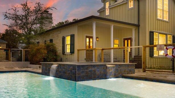 A swimming pool with a waterfall feature, a covered patio, and a house with a stone facade. - Sunset Valley Ranch (Fredericksburg)