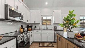 Cozinha moderna com armários brancos, bancada preta, forno de aço inoxidável e micro-ondas. Há uma janela com vista para o exterior, uma planta em vaso e uma fruteira com uvas.