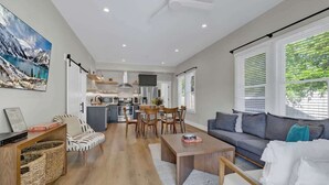 A spacious living area with a sofa, coffee table, and dining table. A kitchen with modern appliances is visible in the background. - The Grove Combo (Fredericksburg)