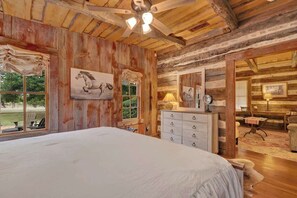 A rustic bedroom with a large bed, wooden walls, a ceiling fan, and a window with a view of the outdoors. - Blakey at Sunset Valley Ranch (Fredericksburg)