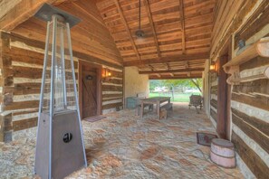 A log cabin interior with a stone floor, a metal heater, wooden furniture, and a view of the outdoors. - Blakey at Sunset Valley Ranch (Fredericksburg)