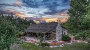 Una cabaña de troncos con chimenea de piedra, rodeada de un césped cuidado y abundante follaje.