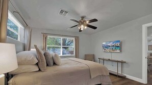 A bedroom with a bed, a ceiling fan, a window with curtains, a bedside table, and a painting on the wall. - Weitz Retreat (Fredericksburg)