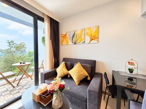 A modern living room with a grey sofa, yellow cushions, a dining table with a metal stand, and a large window with a view of greenery.