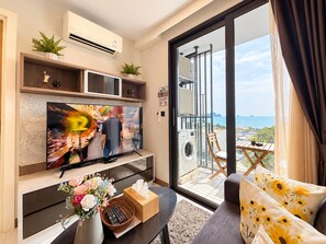 A modern living room with a flat-screen TV, a sofa with floral cushions, a coffee table with a vase of flowers, and a balcony with a washing machine.